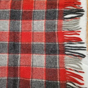 Op de afbeelding: Een close-up van een rood, grijs en zwart geruite deken met gefranjerde randen. De deken heeft een klassiek vierkant patroon met afwisselende kleuren. De franje is rood, grijs en zwart.