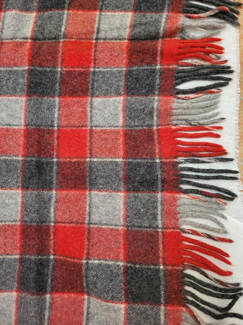 Op de afbeelding: Een close-up van een rood, grijs en zwart geruite deken met gefranjerde randen. De deken heeft een klassiek vierkant patroon met afwisselende kleuren. De franje is rood, grijs en zwart.