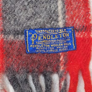 Op de afbeelding: Close-up van een Pendleton wollen sjaal met een rood, grijs en wit ruitpatroon. Een blauw label met gouden tekst luidt "WARRANTED TO BE A PENDLETON" en details over de wol. De sjaal is gemaakt van 100% scheerwol.