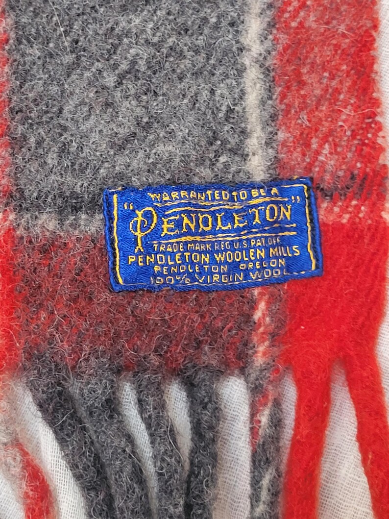 Op de afbeelding: Close-up van een Pendleton wollen sjaal met een rood, grijs en wit ruitpatroon. Een blauw label met gouden tekst luidt "WARRANTED TO BE A PENDLETON" en details over de wol. De sjaal is gemaakt van 100% scheerwol.