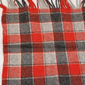 Op de afbeelding: Een close-up van een rood, grijs en zwart geruite deken met franjes. De deken heeft een klassiek ruitpatroon met een mix van rode, grijze en zwarte vierkanten. De franjes zijn een mix van rood en grijs.