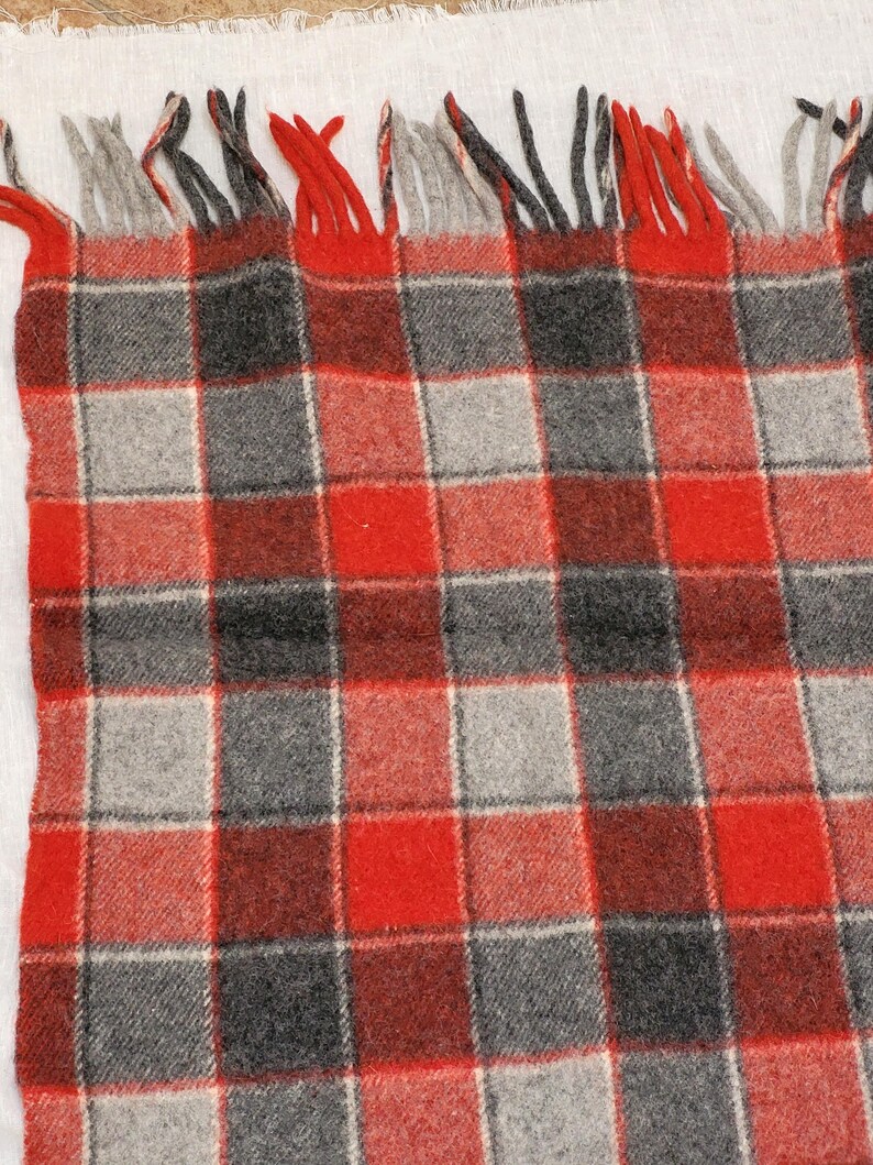 Op de afbeelding: Een close-up van een rood, grijs en zwart geruite deken met franjes. De deken heeft een klassiek ruitpatroon met een mix van rode, grijze en zwarte vierkanten. De franjes zijn een mix van rood en grijs.