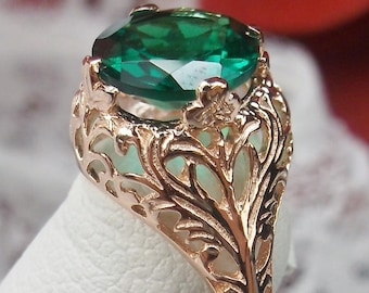 May include: Rose gold ring featuring a large, oval-cut emerald green gemstone. The ring has an ornate, filigree design with floral accents. The intricate metalwork creates a delicate, vintage aesthetic. The ring is displayed on a white surface.