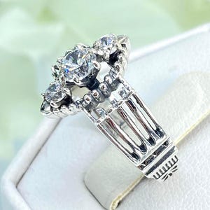 May include: A silver ring featuring a large, clear, round central stone and smaller stones around it. The ring has an intricate, architectural design on the band. The ring is displayed on a white surface.