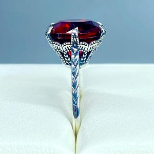 Könnte beinhalten: Ein silberner Ring mit einem großen, oval geschliffenen, tiefroten Edelstein. Der Edelstein ist in einer detaillierten Fassung im Filigranstil gefasst. Das Band des Rings hat ein strukturiertes Design, das seine Vintage-Ästhetik unterstreicht.