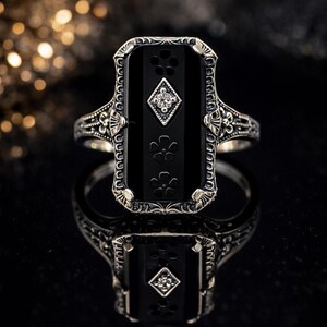 May include: A silver-toned ring with an Art Deco design. The ring features a rectangular black onyx stone with floral and diamond accents. The band has intricate filigree detailing. The ring is displayed on a reflective black surface.