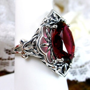 Puede incluir: Un anillo de plata con una piedra preciosa ovalada de color rojo intenso. El anillo tiene un diseño de filigrana ornamentado con detalles florales y de hojas que rodean la piedra. La piedra preciosa está facetada, captando la luz y creando un efecto brillante.