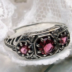Può includere: Un anello d'argento con tre pietre preziose rosa e dettagli intricati. L'anello è posto su uno sfondo di pizzo bianco.