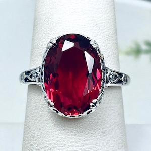 May include: A silver ring featuring an oval, deep red gemstone. The gemstone is the focal point, set in a decorative silver band with intricate filigree detailing. The ring is displayed on a white surface, highlighting its color and design.