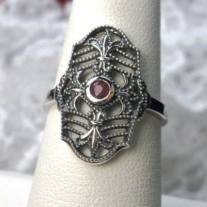 May include: A silver-toned ring with an ornate, filigree design. The ring features a central, round, red gemstone and fleur-de-lis motifs. The intricate metalwork creates a detailed, openwork pattern. The ring is displayed on a white surface.