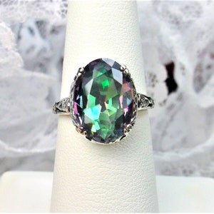 May include: A silver ring with a large oval-shaped gemstone. The gemstone is a mystic topaz, which is a type of quartz that exhibits a rainbow of colors. The ring is set in a delicate filigree design.