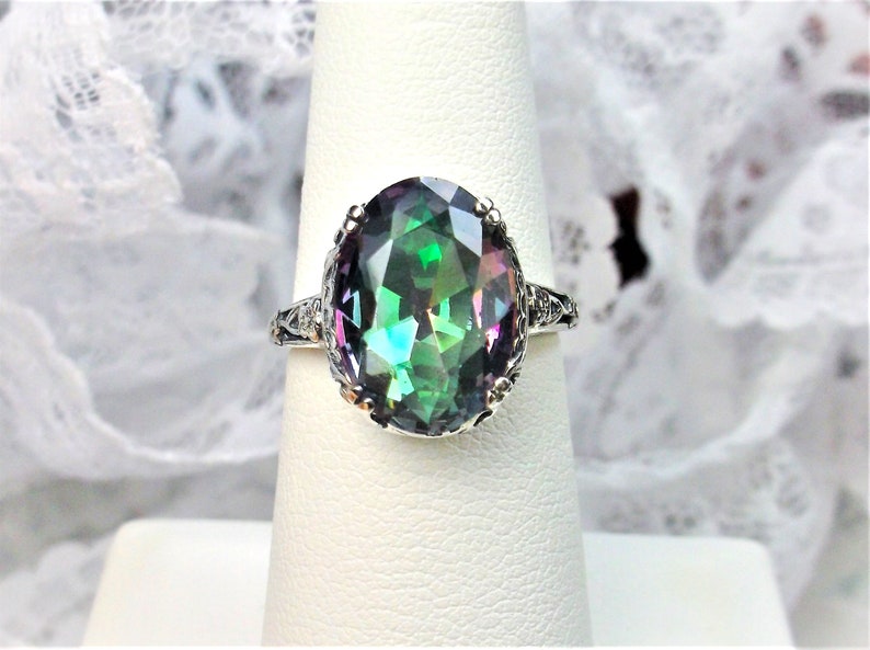 May include: A silver ring with a large oval-shaped gemstone. The gemstone is a mystic topaz, which is a type of quartz that exhibits a rainbow of colors. The ring is set in a delicate filigree design.
