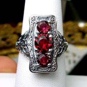 May include: A silver ring with a rectangular setting featuring three red gemstones. The ring has an intricate, floral design.