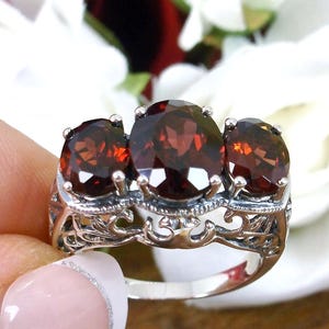 Puede incluir: Un anillo de plata con tres piedras preciosas ovaladas de color rojo intenso. El anillo tiene un diseño de filigrana ornamentado en la banda y alrededor de las piedras. Las piedras están engastadas.