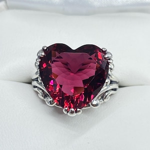 May include: A sterling silver ring featuring a large, heart-shaped, faceted, deep red gemstone. The gemstone is surrounded by a decorative silver setting with ornate details. The ring is set against a white background.