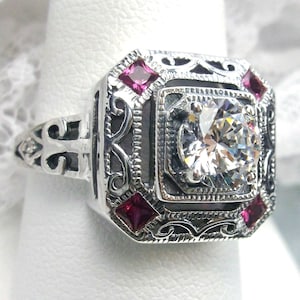 May include: A silver ring with a large, clear, round-cut center stone. The ring features a square setting with filigree detailing and four smaller, square, red gemstones. The ring is on a white background.