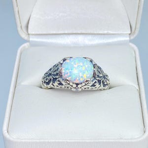 May include: A silver ring with an oval, iridescent opal gemstone. The ring features an ornate, engraved design around the gemstone and a small accent stone on each side. The ring is displayed in a white jewelry box.