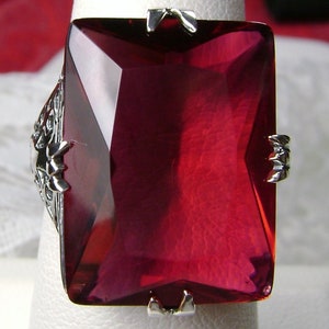 May include: A large, rectangular, ruby-red gemstone ring. The ring features a silver filigree design on the sides and small silver clasps securing the gemstone. The gemstone is faceted, catching the light and displaying a deep red color.