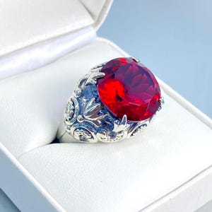 May include: A silver ring with a large, faceted, oval-cut red gemstone. The ring's band features intricate floral and leaf patterns. The ring is displayed in a white jewelry box, set against a light blue background.
