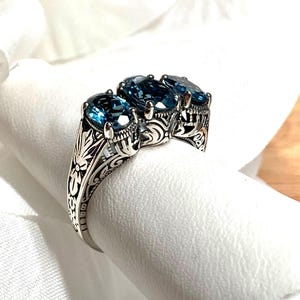 May include: A silver ring with three oval-shaped blue topaz gemstones set in a filigree design.