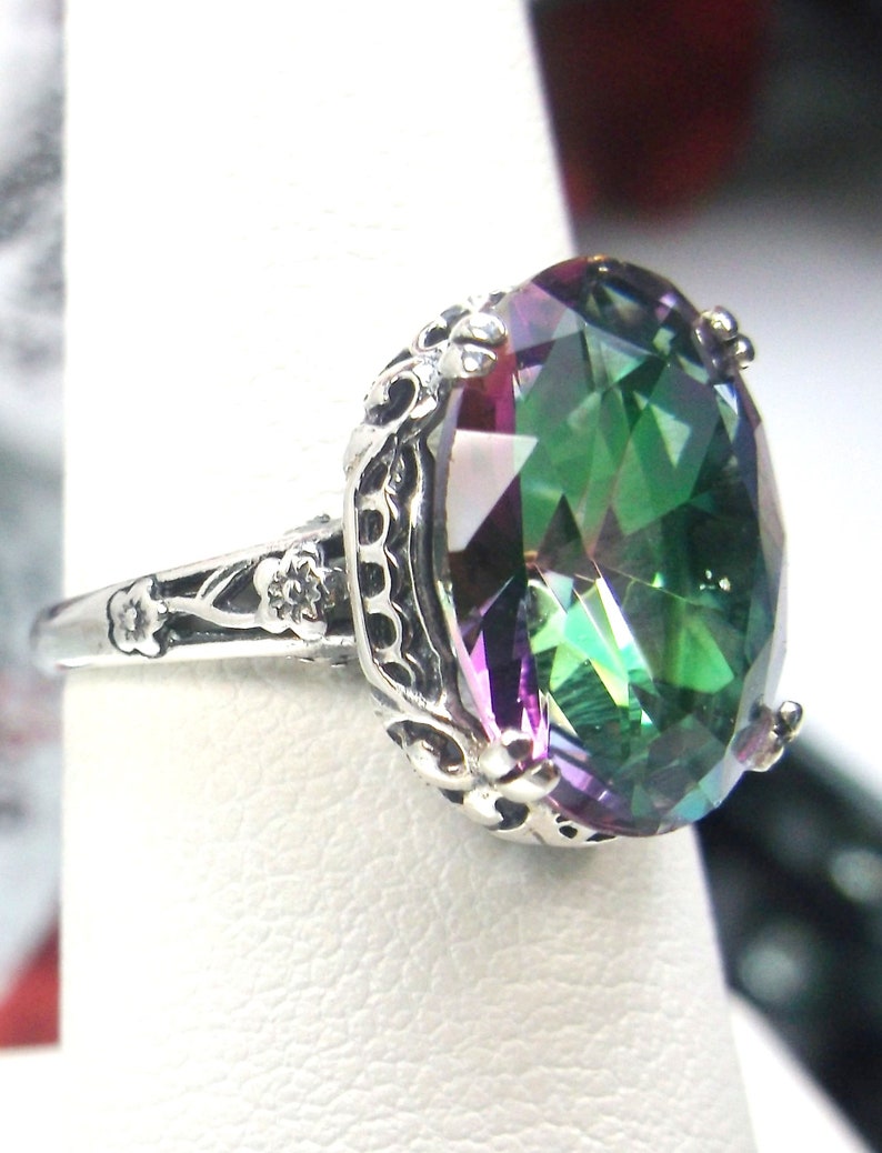 May include: A silver ring with a large, oval-shaped, iridescent gemstone. The gemstone is a mix of green, purple, and blue colors. The ring has a detailed, ornate design.