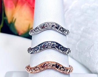 May include: Three ornate, curved rings in silver, black, and rose gold are displayed on a white ring holder. Each ring features a detailed floral and scroll design. The rings are set against a soft, blurred background.