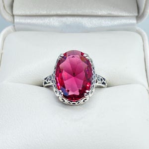 May include: A silver ring with a large, oval-cut, ruby-red gemstone. The ring has intricate filigree detailing on the band and around the gemstone setting. The ring is displayed in a white jewelry box.