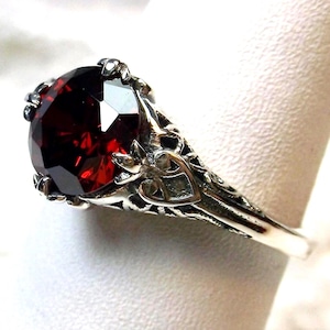 May include: A silver ring with a large, red gemstone set in a detailed, ornate design. The ring has a filigree pattern and a heart-shaped design on the band.