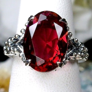 Pode incluir: Um anel de prata com uma grande pedra vermelha de forma oval. O anel tem um design detalhado e ornamentado.