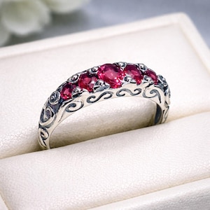 May include: A silver ring featuring five round, deep red gemstones set in a row. The band is crafted with intricate, dark-oxidized scrollwork patterns that wrap around the sides, providing a contrast to the polished silver and the vibrant red stones.