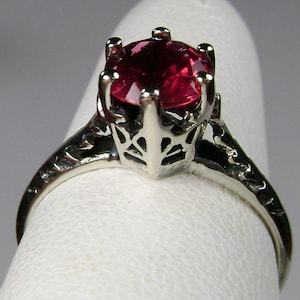 May include: A silver ring with a red gemstone set in a crown-like design. The ring is on a white display stand.