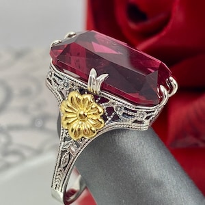 May include: A silver ring with a large, dark red gemstone and a gold flower design. The ring is set on a gray surface.