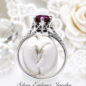 May include: A silver ring with an ornate filigree design and a large, oval, deep red gemstone. The ring is displayed on a white surface, with the words "Silver Embrace Jewelry" at the bottom.