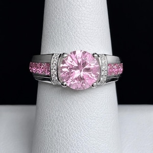 May include: A silver-toned ring featuring a large, round, faceted pink center stone held by four prongs. The band is accented with a row of smaller, round pink stones on each side, bordered by tiny, clear, sparkling stones set in a channel.