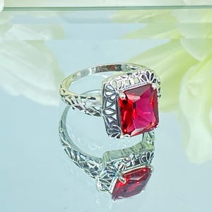 May include: A sterling silver ring with a large, square, red gemstone. The ring features an ornate, filigree design around the gemstone and on the band. The ring is displayed on a reflective surface, showcasing its intricate details and vibrant color.