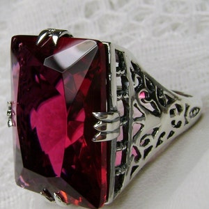 May include: A silver-toned ring featuring a large, rectangular, deep red gemstone. The ring has an ornate, openwork design on the band and decorative metalwork around the gemstone, creating a vintage aesthetic.