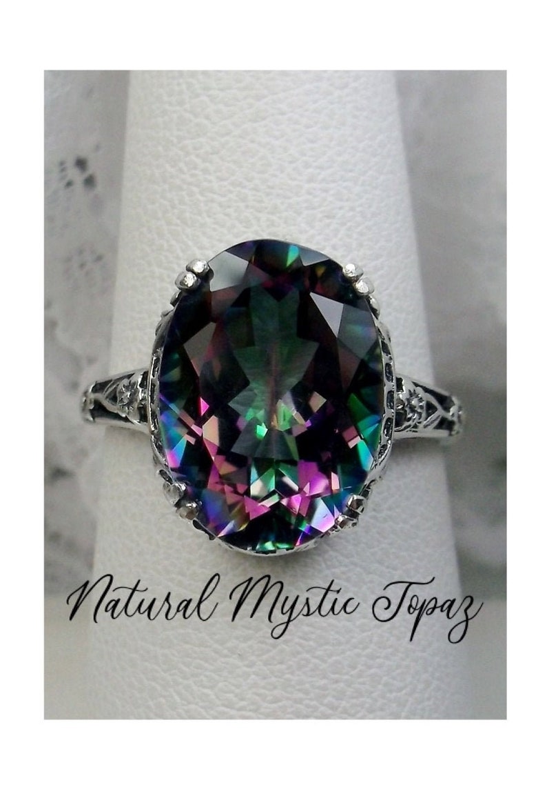 May include: A silver ring with a large oval-shaped mystic topaz gemstone. The gemstone is iridescent and displays a rainbow of colors, including green, blue, purple, and pink. The ring has a delicate filigree design.