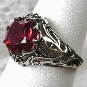 May include: A silver ring featuring a large, faceted, deep red gemstone. The ring has an ornate, swirling design around the stone and along the band, with intricate details. The gemstone is the focal point, catching the light and reflecting a vibrant color.