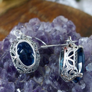 May include: A pair of silver earrings with a dark blue gemstone set in an intricate filigree design. The earrings are displayed on a bed of purple amethyst crystals.