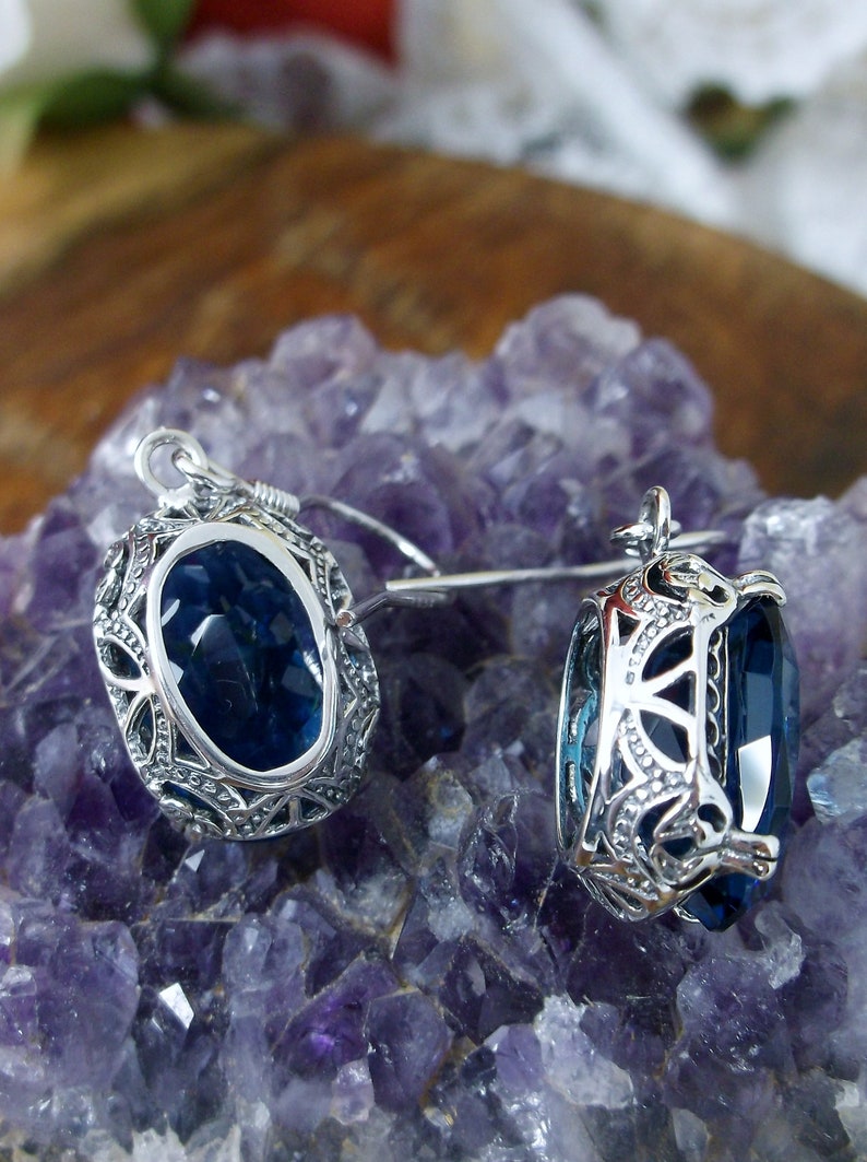 May include: A pair of silver earrings with a dark blue gemstone set in an intricate filigree design. The earrings are displayed on a bed of purple amethyst crystals.
