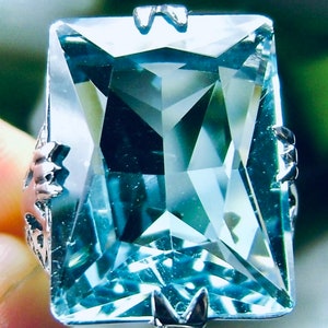 May include: A large, rectangular gemstone ring with a light blue hue. The gemstone is set in a silver-colored metal band with decorative filigree on the sides. The ring's design features a geometric, faceted cut.