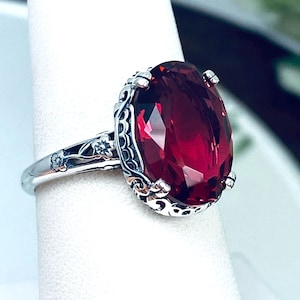 May include: A silver ring featuring an oval, faceted, deep red gemstone. The ring has an ornate, filigree design around the gemstone and on the band, with small floral accents. The ring is displayed on a white surface.