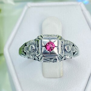 May include: A silver ring with a square center setting featuring a small, round, pink gemstone. The ring has intricate filigree detailing on the band and around the gemstone. The ring is displayed on a white stand.