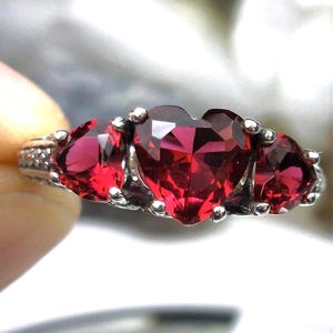May include: A silver ring with three heart-shaped red gemstones. The ring is set with a large heart-shaped gemstone in the center, flanked by two smaller heart-shaped gemstones.