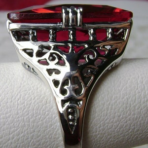 May include: A silver ring featuring a large, rectangular, faceted red gemstone. The ring has an ornate, openwork design on the sides and a detailed setting around the gemstone. The ring is shown on a white surface.