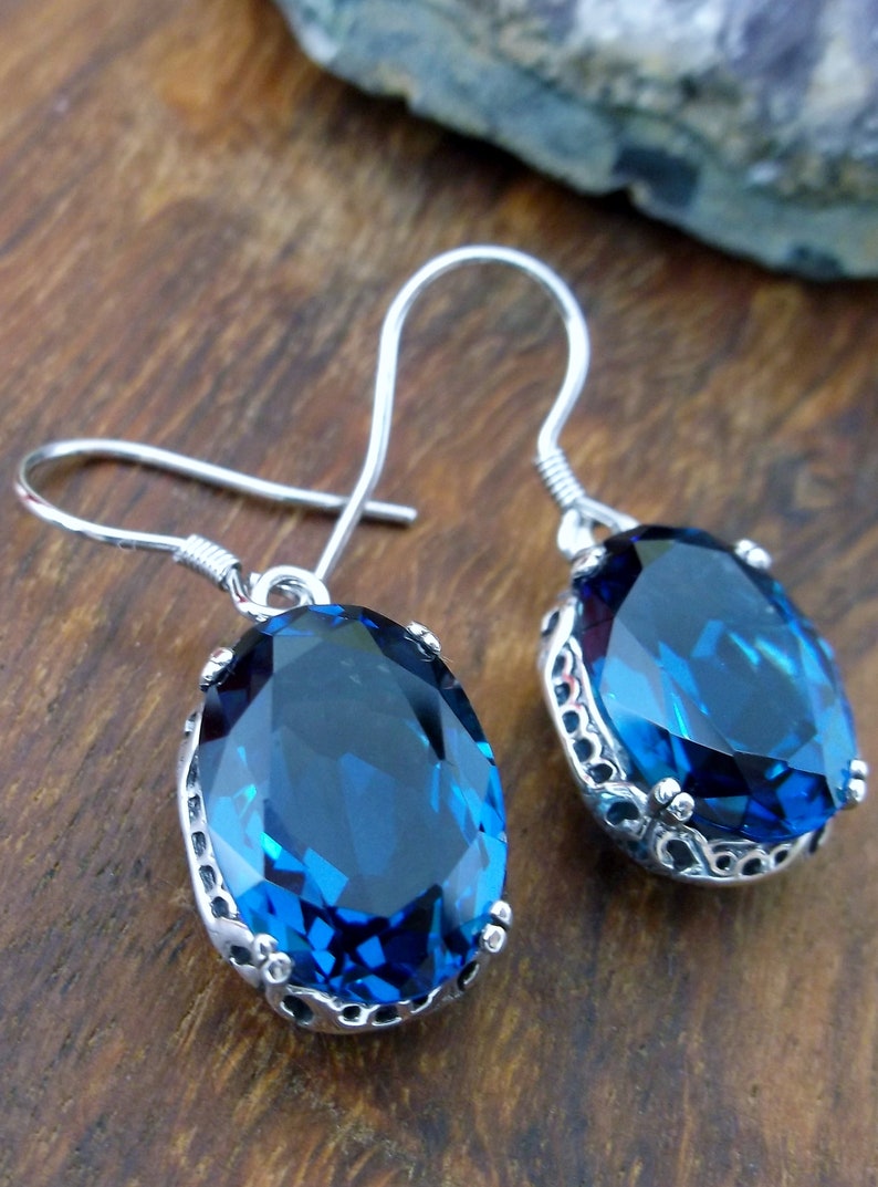 May include: A pair of silver earrings with blue gemstone oval drops. The earrings have a delicate filigree design around the stones.