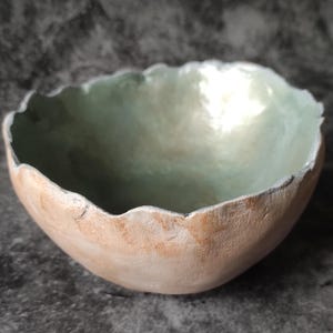 May include: A handmade, organic-shaped bowl with a rough, textured exterior in a light peach color. The interior is a soft, pearlescent green. The rim has a scalloped edge, adding to its unique, artistic appeal. The bowl is likely used for decorative purposes.