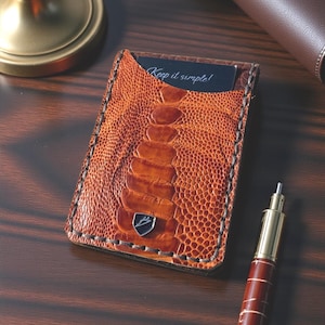May include: A brown leather cardholder with an ostrich leg pattern. The cardholder has a stitched border and a small emblem. A card with the text "Keep it simple!" is visible. A pen is in the foreground.