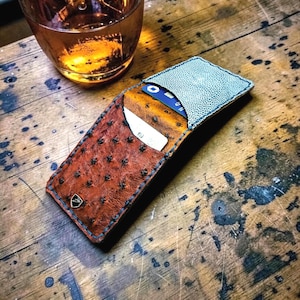 May include: A brown leather cardholder with a textured pattern and blue stitching, holding credit cards. The cardholder is open, revealing the interior. A glass of amber liquid sits nearby on a wooden surface.