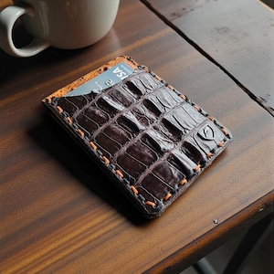 May include: A brown leather wallet with a crocodile skin pattern, orange stitching, and a card peeking out. The wallet is on a wooden surface next to a cup of coffee with latte art.